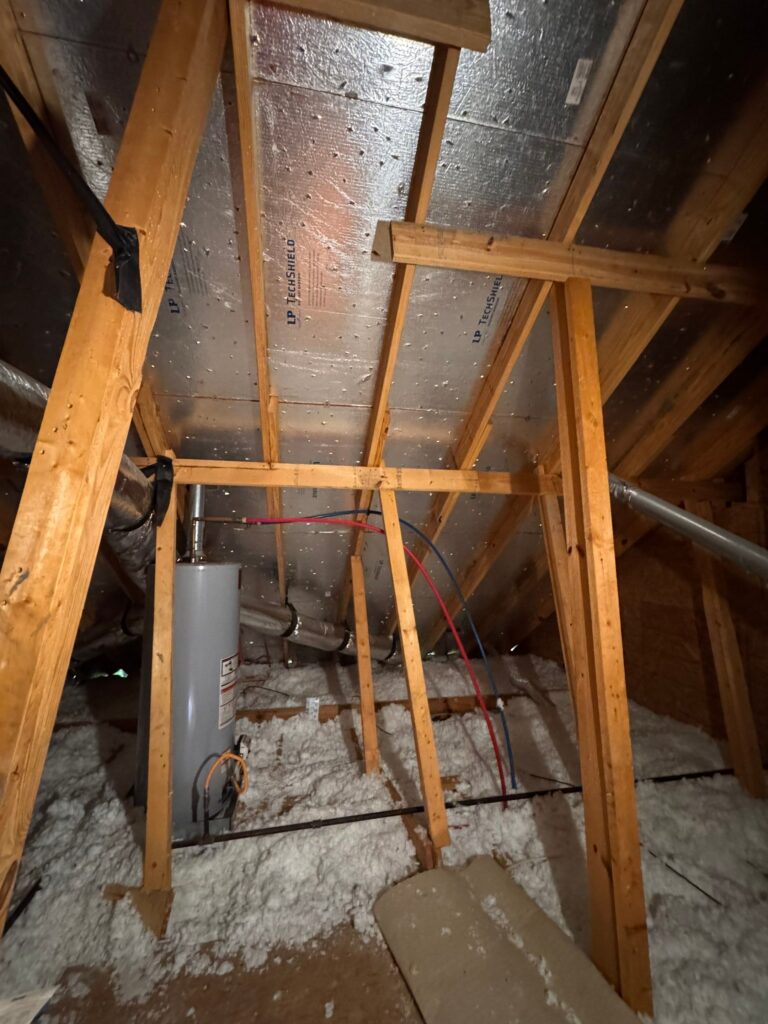 This view shows the interior of an attic with a water heater installed, featuring radiant barrier sheathing on the underside of the roof deck and blown-in insulation covering the floor.