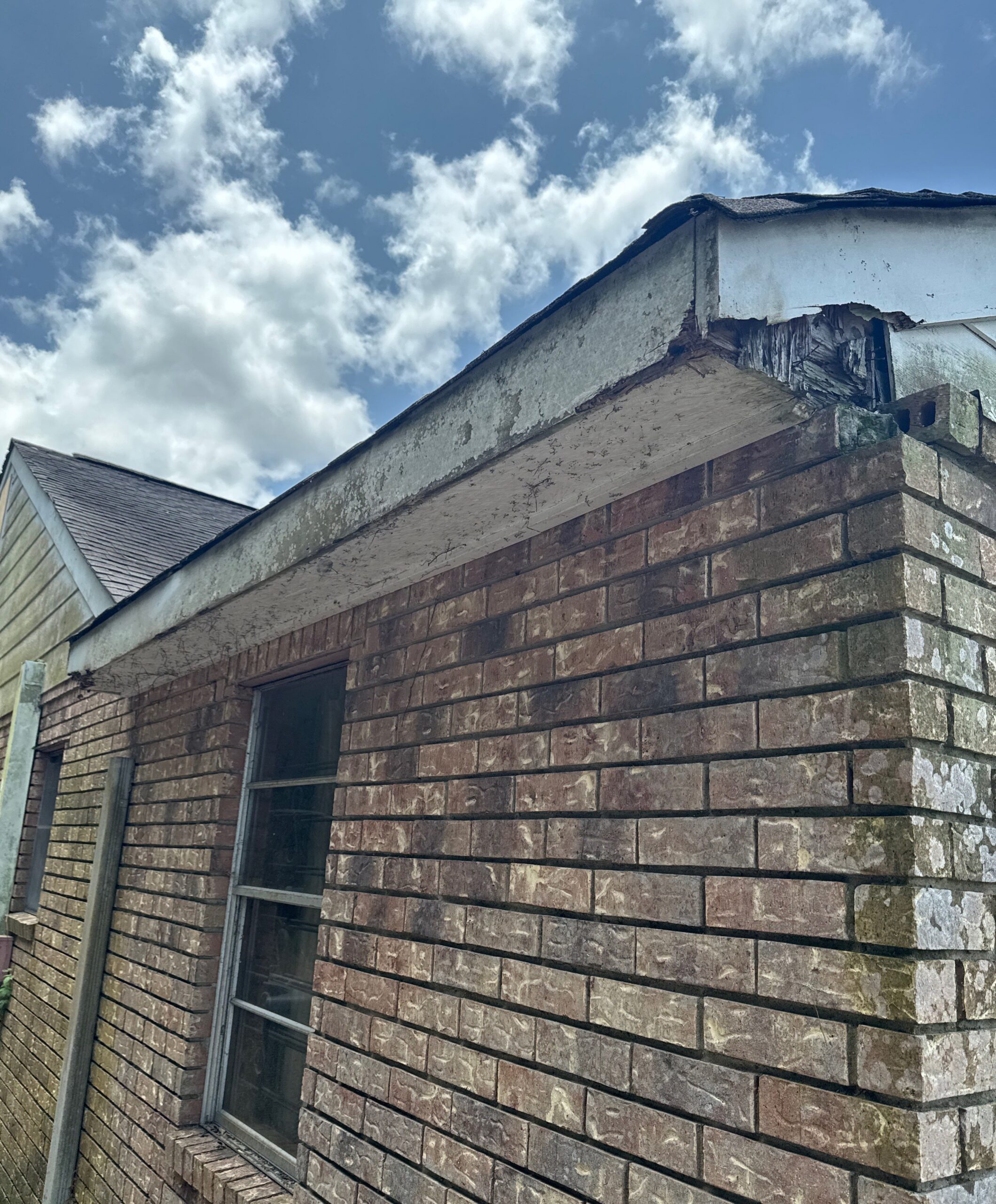 A close-up view of a brick residential structure showing significant wood rot on the fascia board and soffit area near the roofline. The damaged wood reveals exposed structure where the roof edge meets the brick masonry.