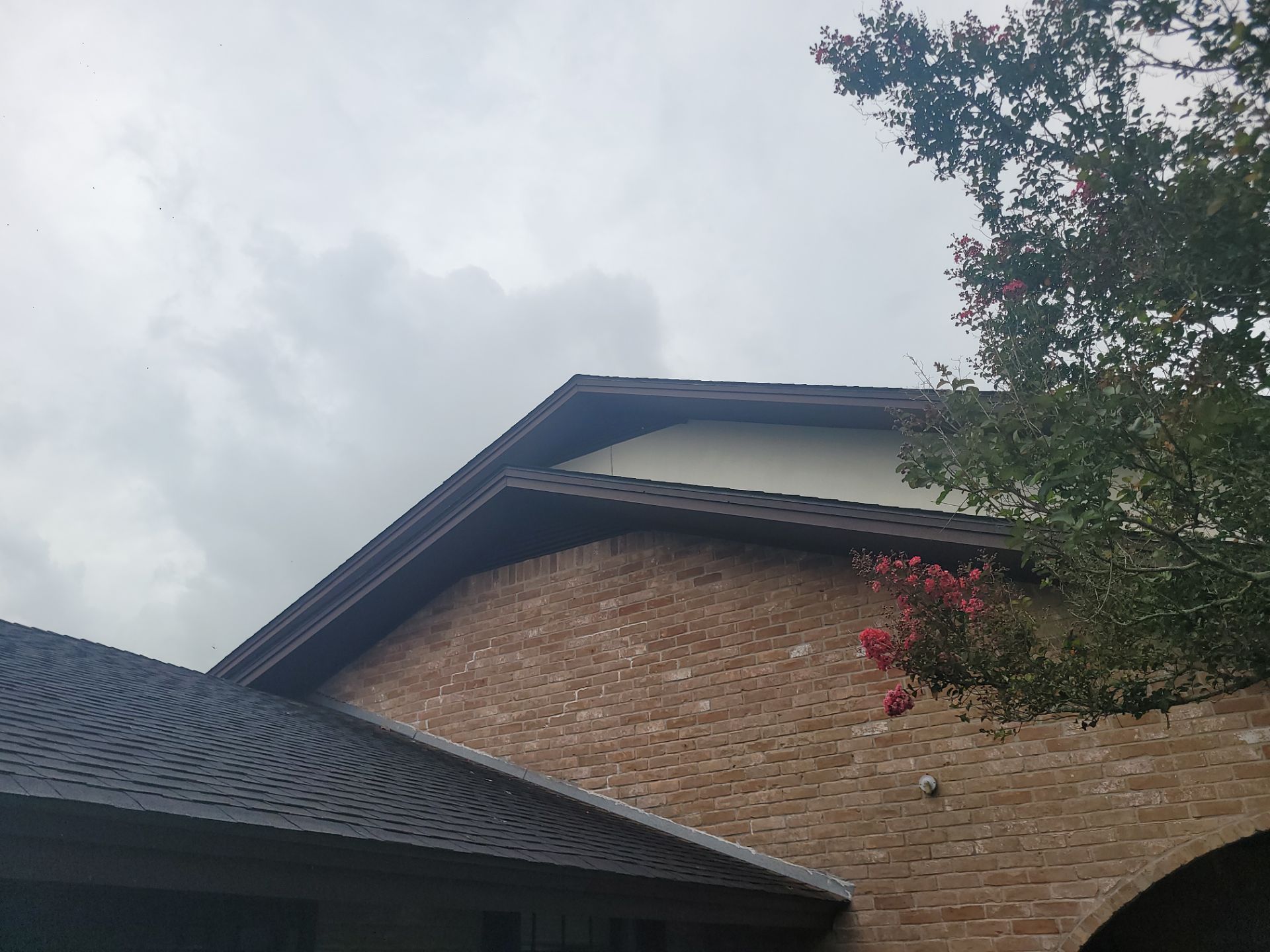 A view of a residential brick house exterior showing a multi-level gable roofline with asphalt shingles and a cream-colored soffit panel. A crepe myrtle tree with pink flowers is visible on the right side.