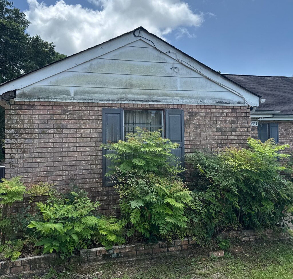 A single-story brick house with dark gray shutters features a gable end with soiled, mildew-covered siding that requires professional cleaning. The exterior siding near the roofline shows significant discoloration and organic growth buildup.