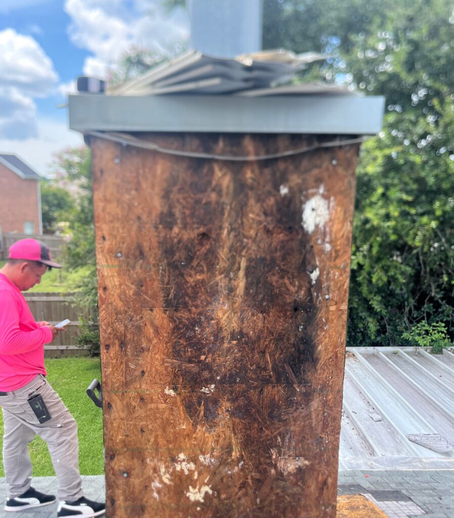 A contractor stands on a residential roof inspecting the plywood sheathing of a chimney chase that has been stripped of its exterior siding, revealing dark signs of moisture damage.
