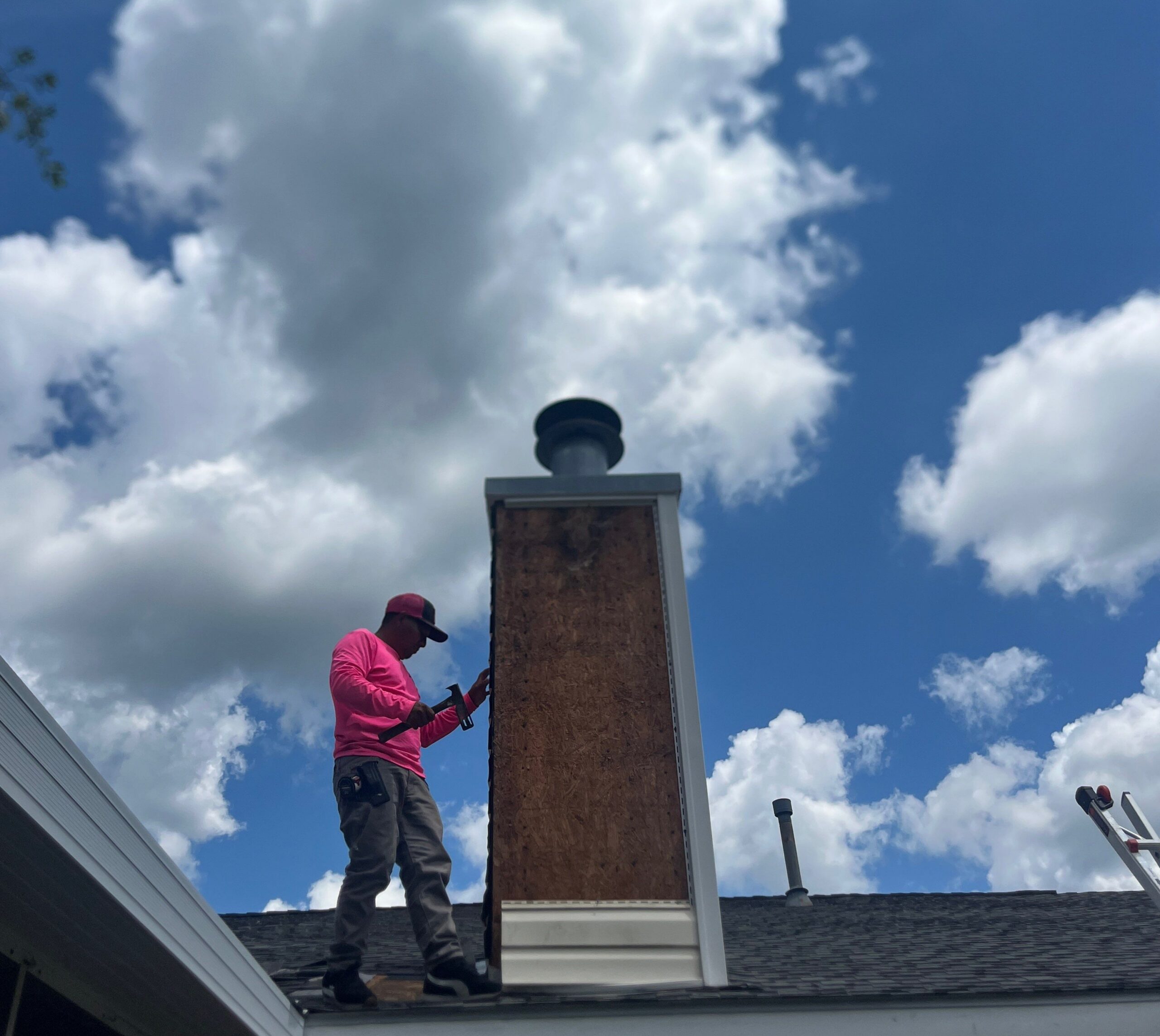 A contractor in a pink shirt is working on the exterior of a chimney on a residential roof. The chimney has exposed plywood sheathing where the siding has been removed, and the worker is using a hammer to perform repairs.