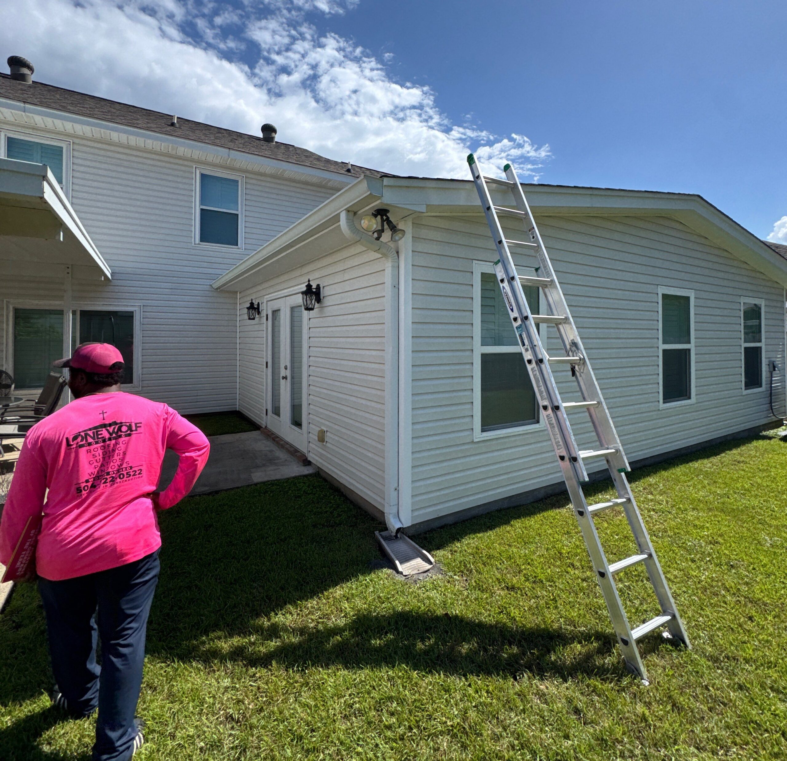 A contractor wearing a branded pink shirt stands in a backyard near a house where an extension ladder is leaning against the side of the roof, likely preparing for an inspection or maintenance.