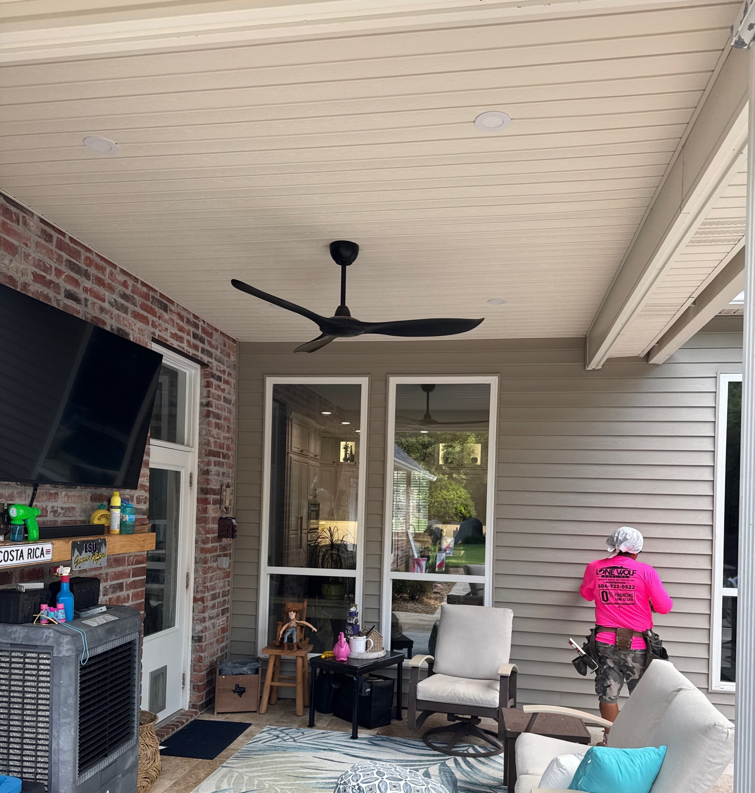 A contractor wearing a pink uniform is installing siding on the exterior wall of a covered patio. The workspace features patio furniture, an outdoor rug, and a large television mounted on a brick wall.