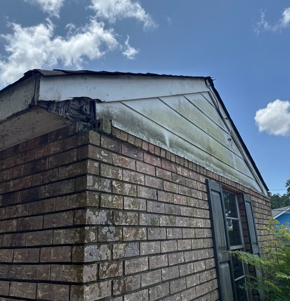 A close-up view shows significant rot and damage to the fascia board and underlying wood framing at the eave of a brick home. The wooden siding on the gable end also shows signs of water staining and organic growth.