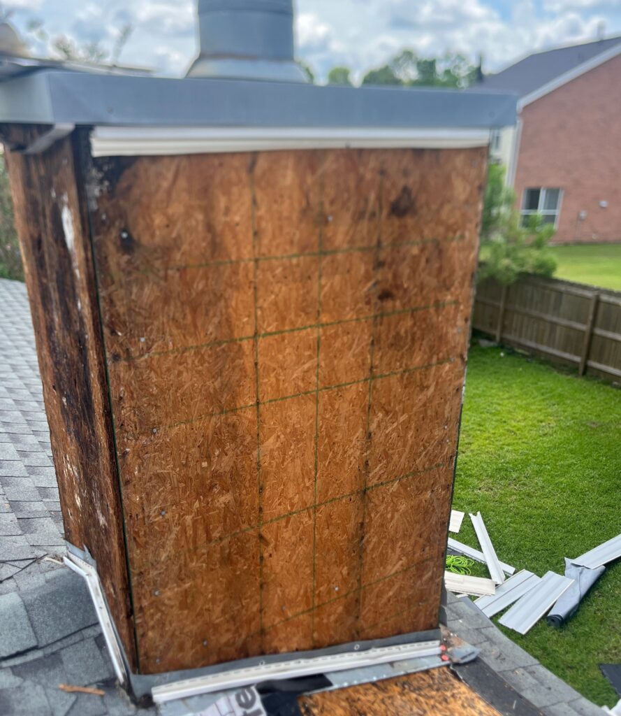 A residential chimney on a shingled roof has its siding removed, exposing the oriented strand board sheathing. The wood appears to have some moisture damage or staining near the corner.