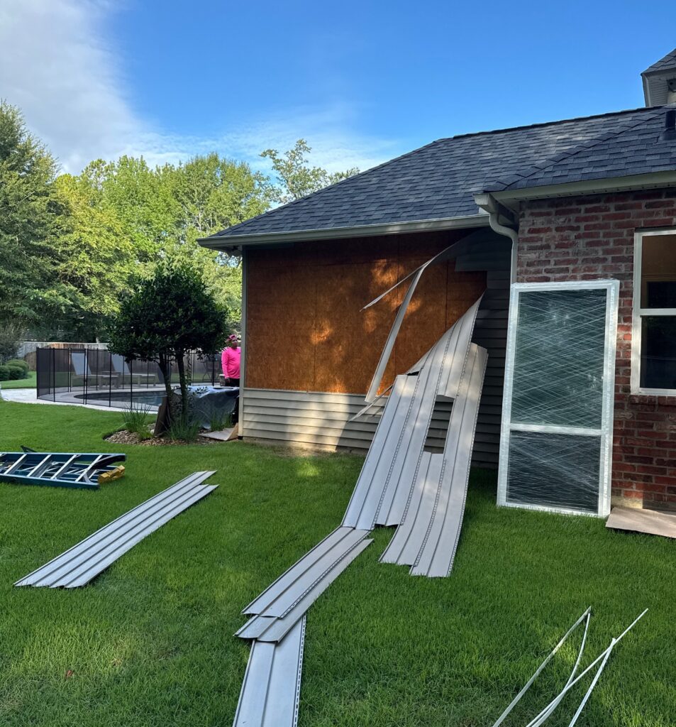 A contractor is working on exterior siding repairs, with removed siding panels lying on the grass and a new window unit leaning against the brick wall.