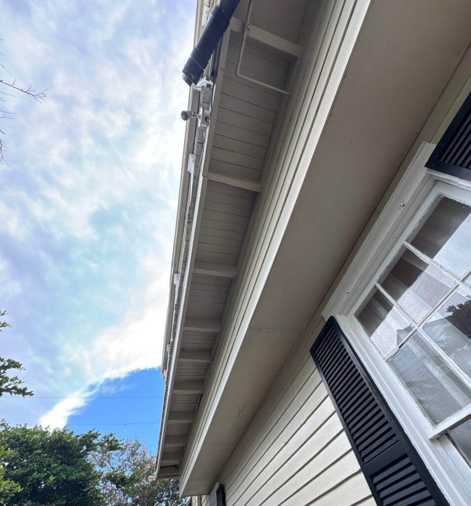 A low-angle view of a house exterior showing the soffit, fascia, and siding. A security camera is mounted under the roof eave near the window shutters.
