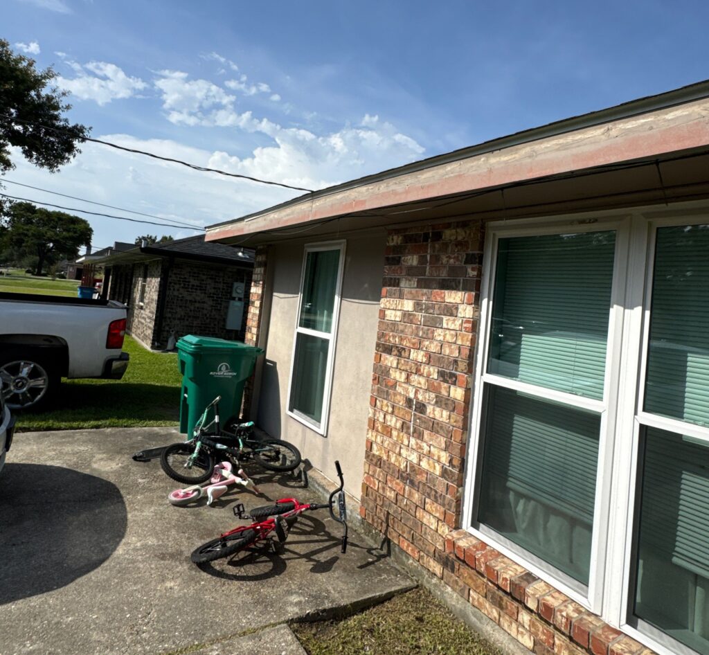 The photograph displays a section of a residential exterior with noticeable deterioration and weathering on the wood fascia board along the roofline. Below, several bicycles and a waste bin are situated on the concrete driveway adjacent to the brick and stucco wall.