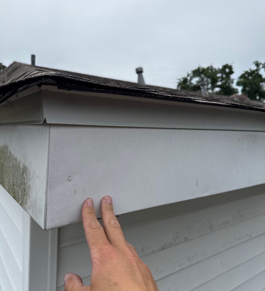 A close-up view of a hand touching the white aluminum fascia covering on a residential roof edge, showing some discoloration and dirt buildup on the corner.