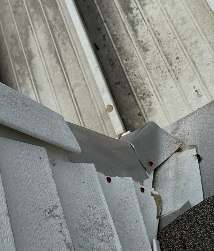 A close-up view of a roof-to-wall transition showing metal flashing and white vinyl siding. The flashing appears to be improperly integrated, showing exposed gaps, rusted fasteners, and potential water intrusion points where the roofing meets the siding.