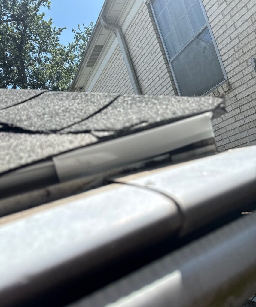 A close-up view showing a gap between the roof edge and the gutter system. The asphalt shingles overhang the gutter, but there is visible separation from the fascia.
