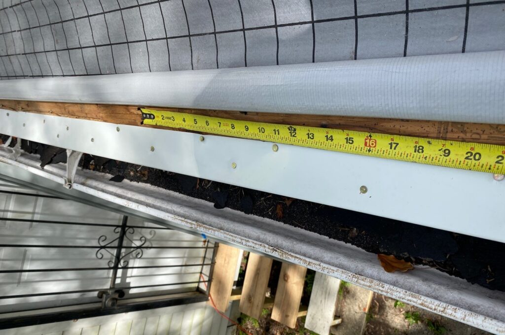 A close-up view showing a tape measure positioned along a roof edge where a gutter meets a fascia board, revealing debris buildup within the gutter system and the underlying roofing felt.