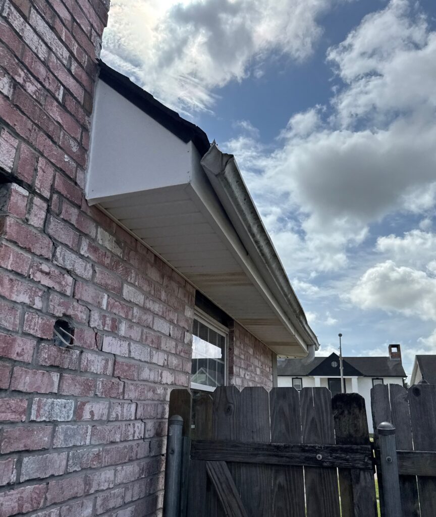 An exterior inspection photo showing the gutter, soffit, and fascia system along the edge of a brick home. A portion of the gutter appears detached or damaged at the corner, with visible weathering on the building materials.