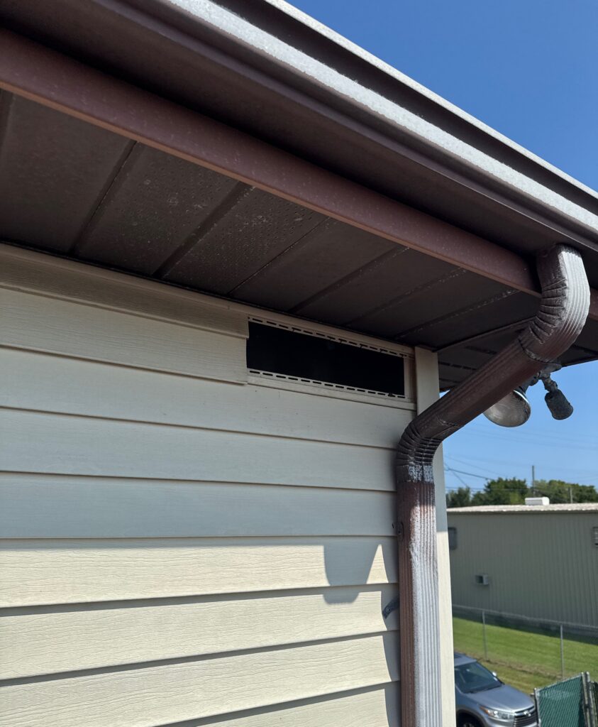 A section of the soffit vent cover is missing, leaving an open rectangular hole in the eaves of the building near the downspout.
