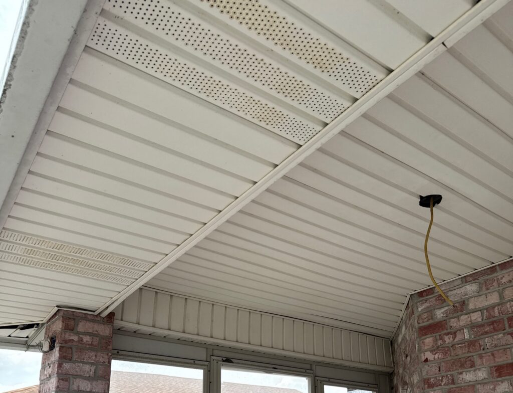 An exterior view of a covered patio featuring white vinyl soffits and brick columns. A yellow electrical wire is hanging through a hole in the ceiling, indicating preparation for a light fixture or ceiling fan installation.