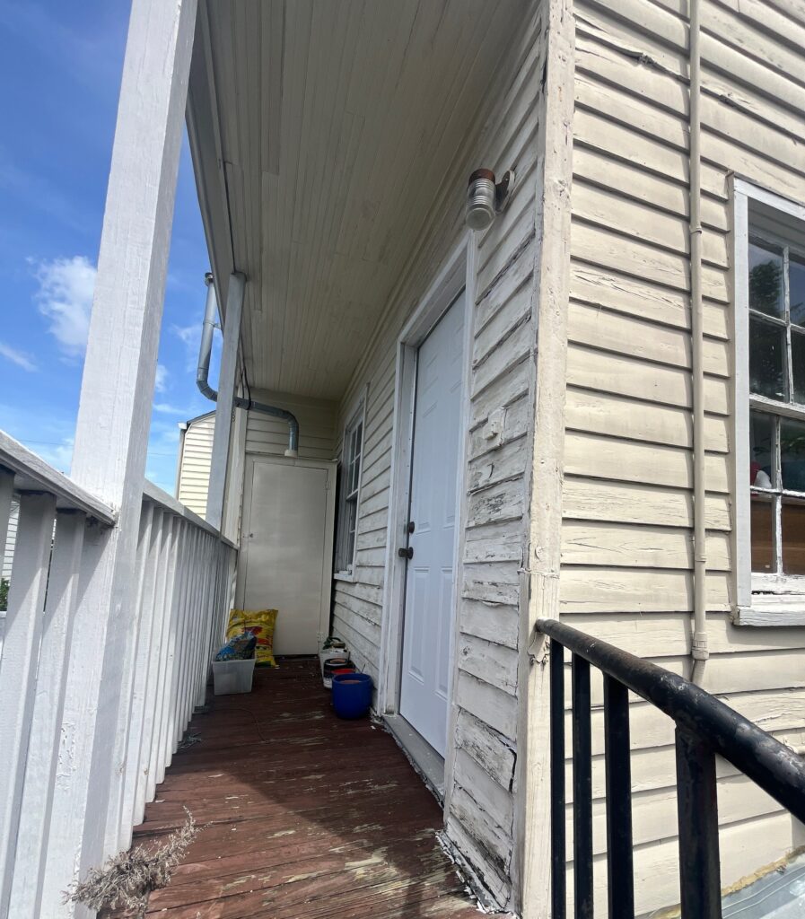 An exterior view of a porch showing peeling white painted wood siding on a home. The deck floorboards appear weathered, and there is a white door and window along the side wall.