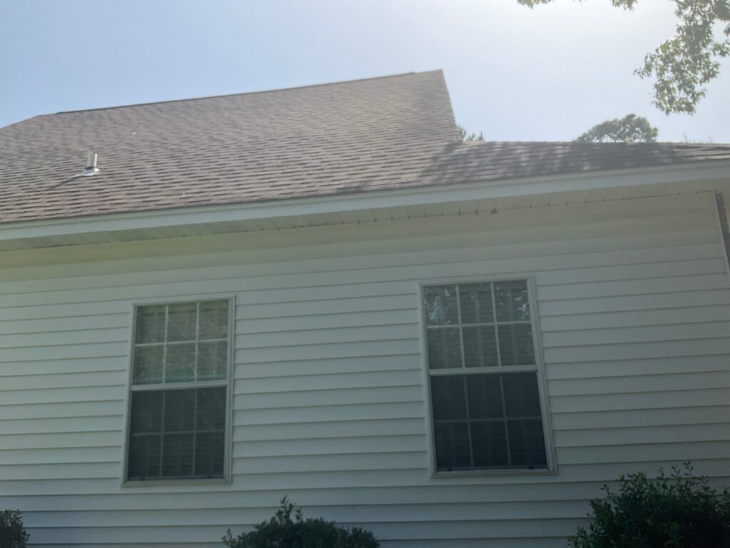 A view of the side of a residential house featuring gray asphalt shingles on the roof and white horizontal vinyl siding. There is an exposed plumbing vent pipe visible on the roof slope above two windows.
