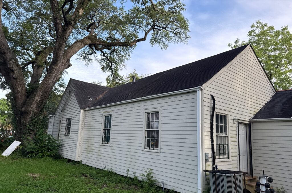 A view of the side profile of a white-sided residential building with dark asphalt shingles. The image shows the exterior walls and roof structure, including windows, an air conditioning unit, and a large oak tree nearby.