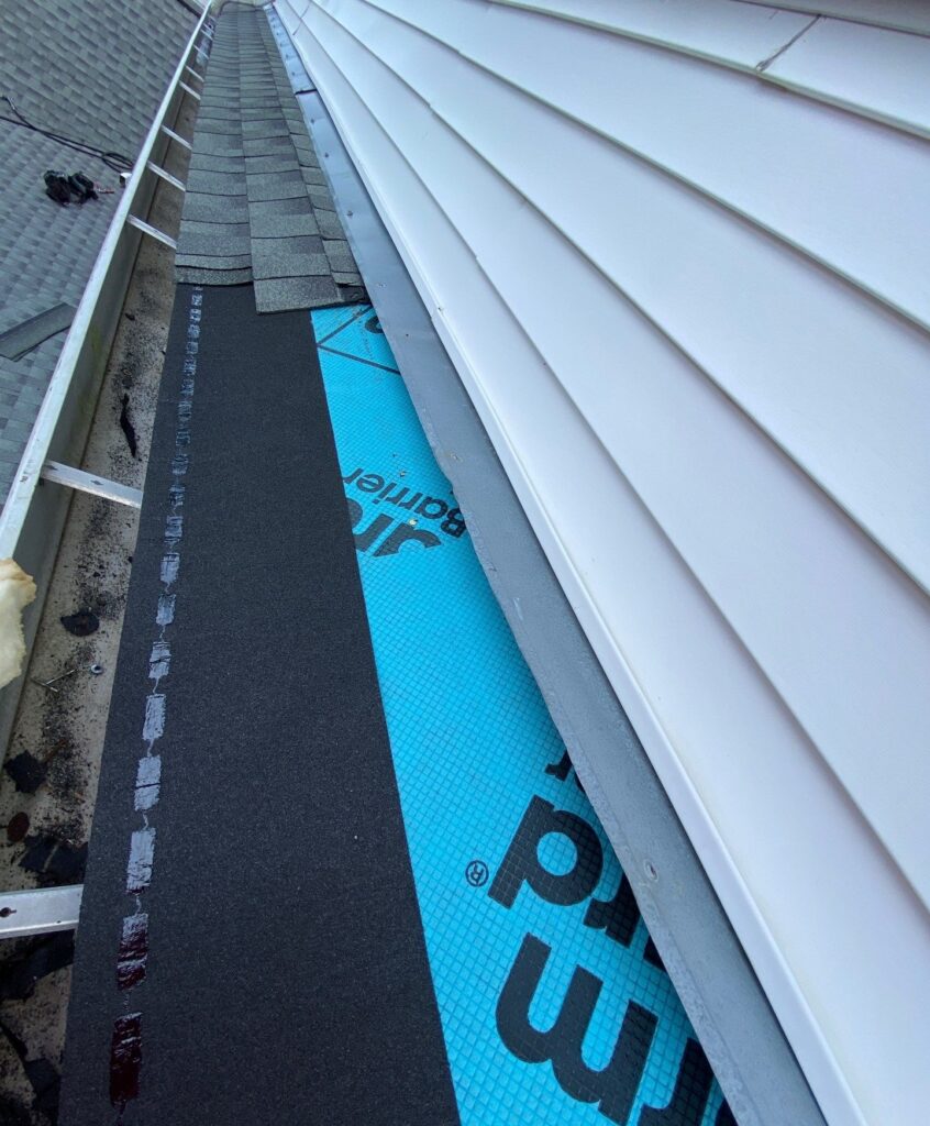 This photo shows a roof repair in progress where new underlayment and shingles are being installed along the sidewall of a house. The image highlights the transition between the blue synthetic underlayment, the black roofing felt, and the existing vinyl siding.