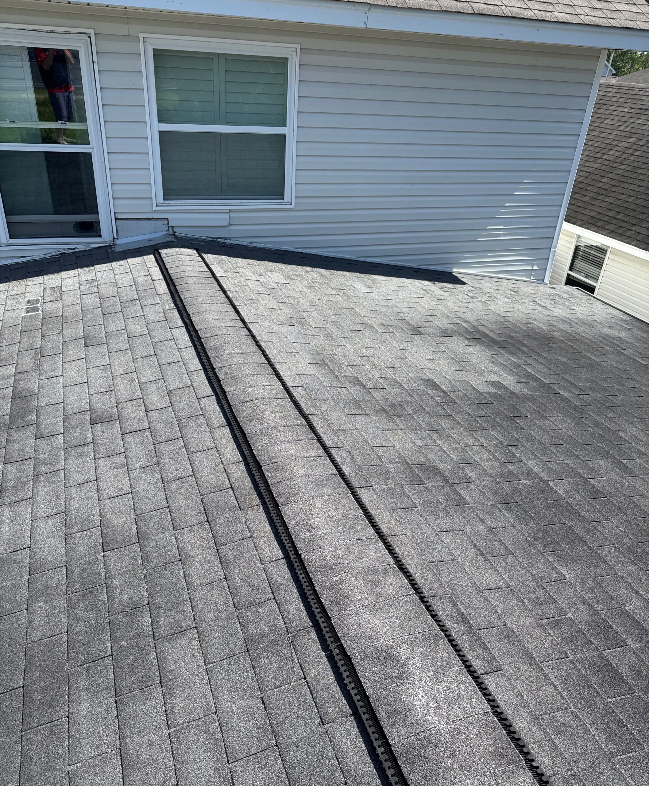 A close-up view of a shingle roof showing an installed ridge vent running along the hip of the roof. The installation appears to be partially complete, with the ridge cap shingles yet to be applied over the venting system.