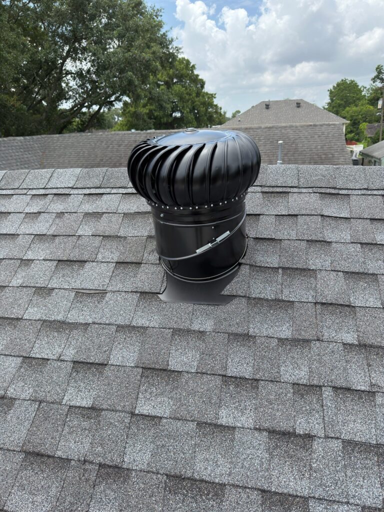 A newly installed black metal turbine roof vent is mounted on a gray asphalt shingle roof. The flashing around the base is integrated into the shingle layers to provide a weather-tight seal.