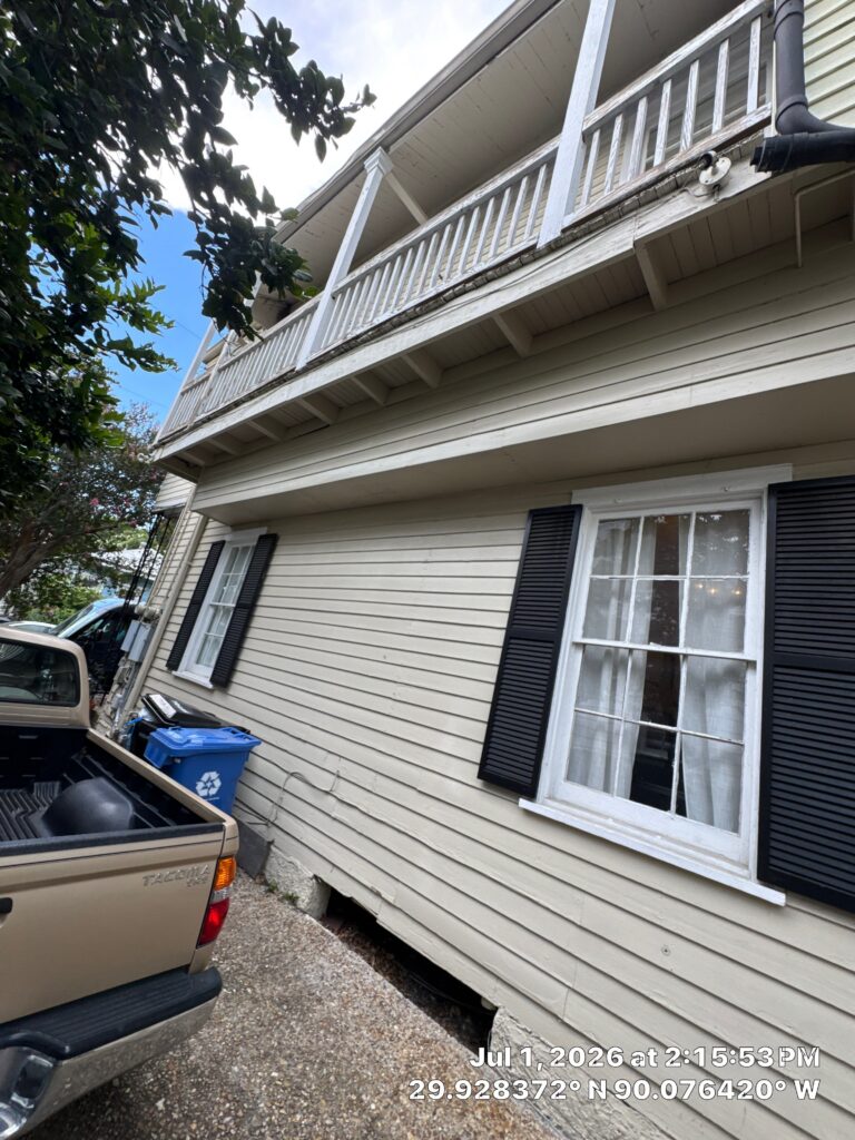A view of the exterior wall of a building showing horizontal lap siding, a second-story balcony with railings, and two windows with black shutters. A Toyota Tacoma is parked in the foreground next to the house.