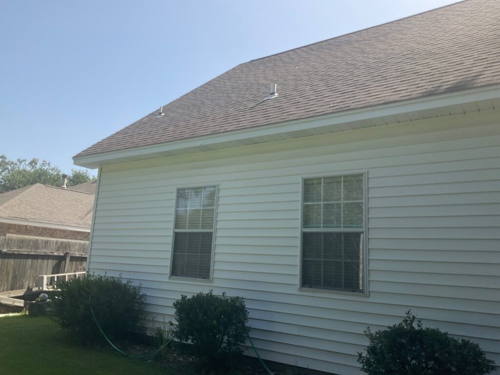 A view of the side of a house with white horizontal vinyl siding and grey asphalt shingle roofing. There are two windows on the wall and two plumbing vents protruding from the roof slope.