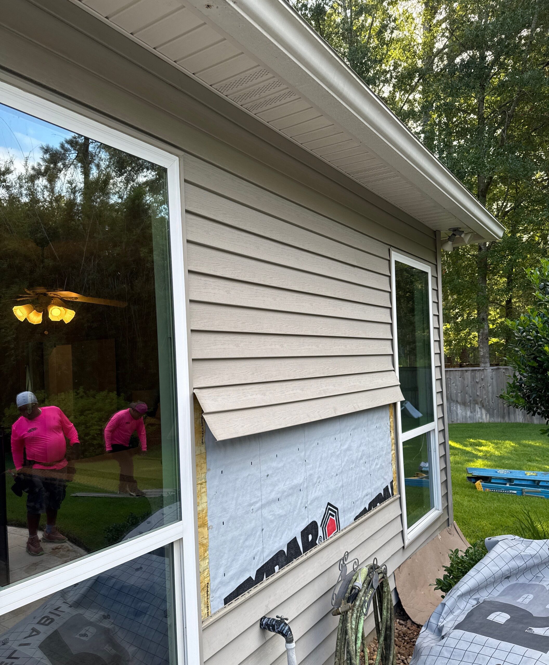 A contractor in a bright pink shirt is installing beige horizontal vinyl siding on a residential home. A section of the wall is exposed, showing the underlying weather barrier wrap.