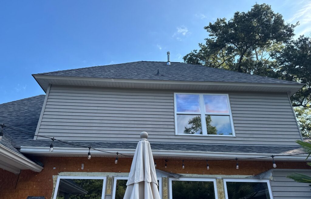 A residential exterior showing ongoing construction where the lower portion of the wall has had siding removed, exposing the sheathing above patio doors. A closed patio umbrella stands in the foreground, and the upper level features gray lap siding and an asphalt shingle roof.