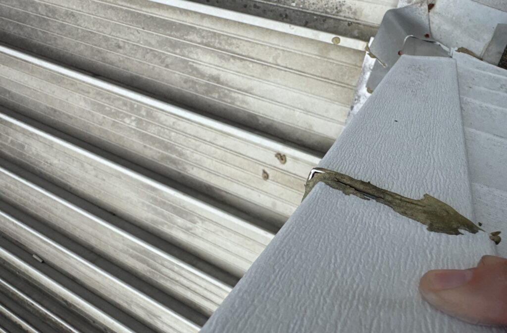 A close-up view of a metal roof fascia trim piece showing a gap in the joint filled with aged, deteriorating exterior sealant. A finger is holding the edge of the material to expose the separation.