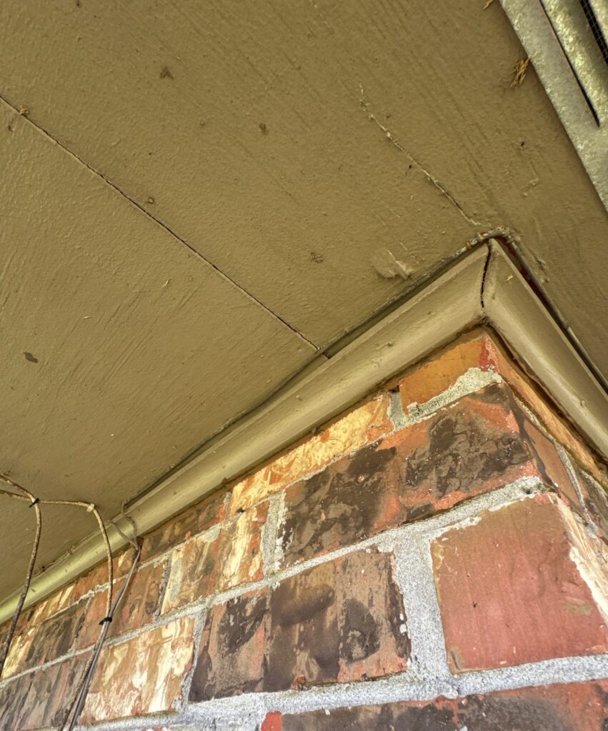 A view showing a gap between the wooden soffit and the brick wall where the fascia trim meets the roofline. There is visible separation and cracking along the exterior corner joint.