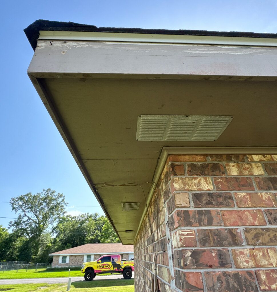 A view of the house soffit highlighting two metal intake vents installed under the roof overhang. The image includes a view of the brick exterior wall and a Lone Wolf contractor truck parked in the background.